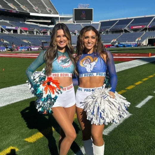 Two cheerleaders in blue and white uniforms standing on a football field with stadium seats in the background.